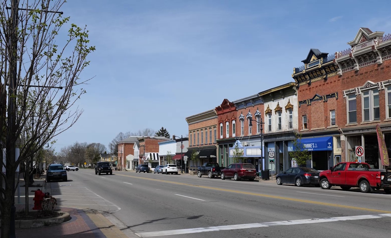 Downtown main street with historic brick storefronts, parked cars along the curb and light traffic on a wide road under a clear sky.