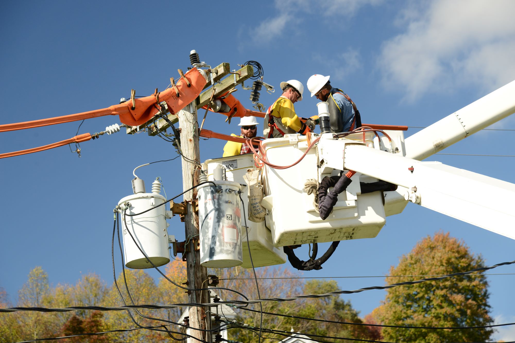Two utility lineworkers in a bucket lift repair electrical equipment on a wooden power pole, surrounded by power lines and transformers against a blue sky and autumn trees. Another lineworker is positioned in a second bucket truck behind them.