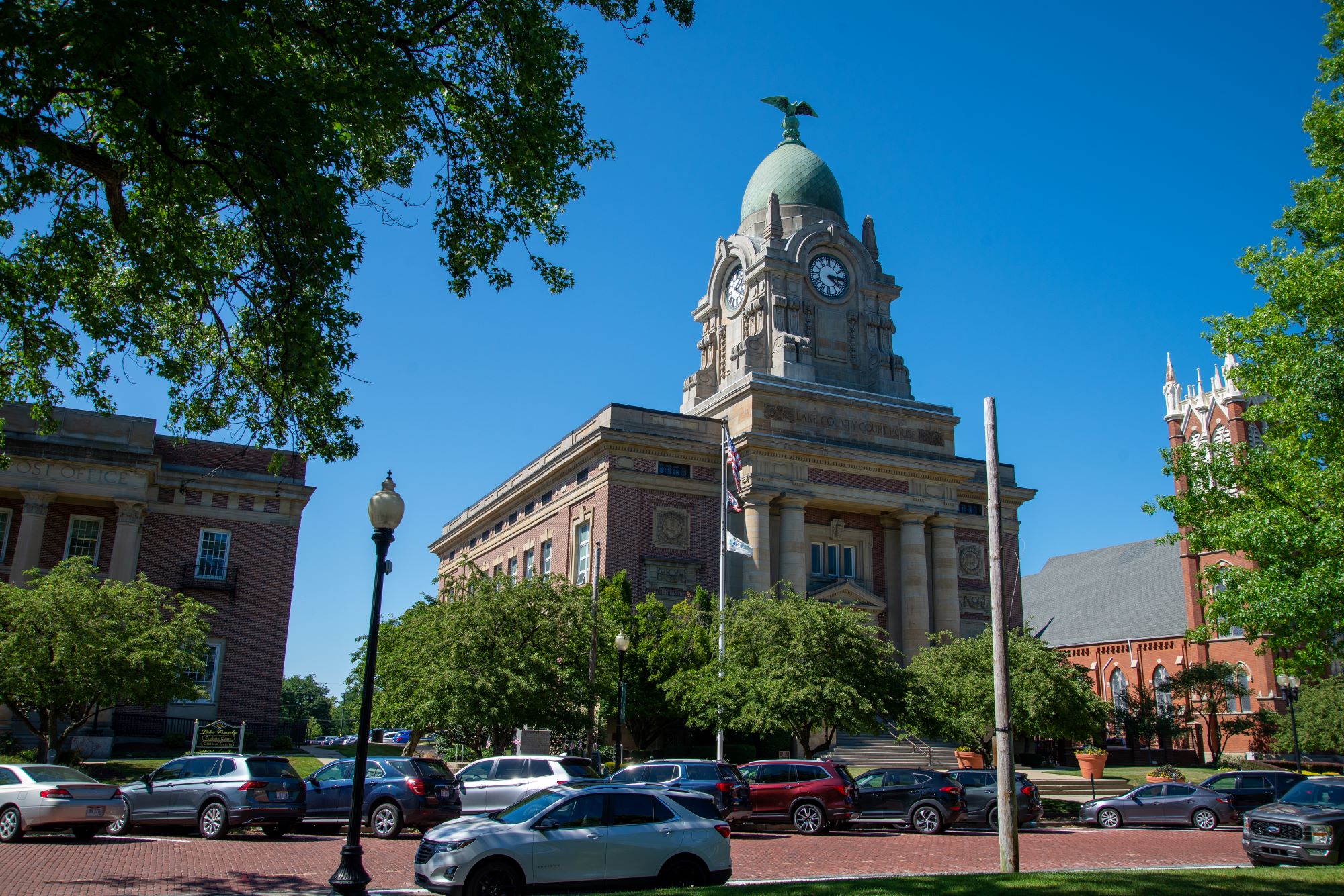 Historic courthouse with a domed clock tower and classical stone details, set among trees with parked cars along a brick street under a clear blue sky.