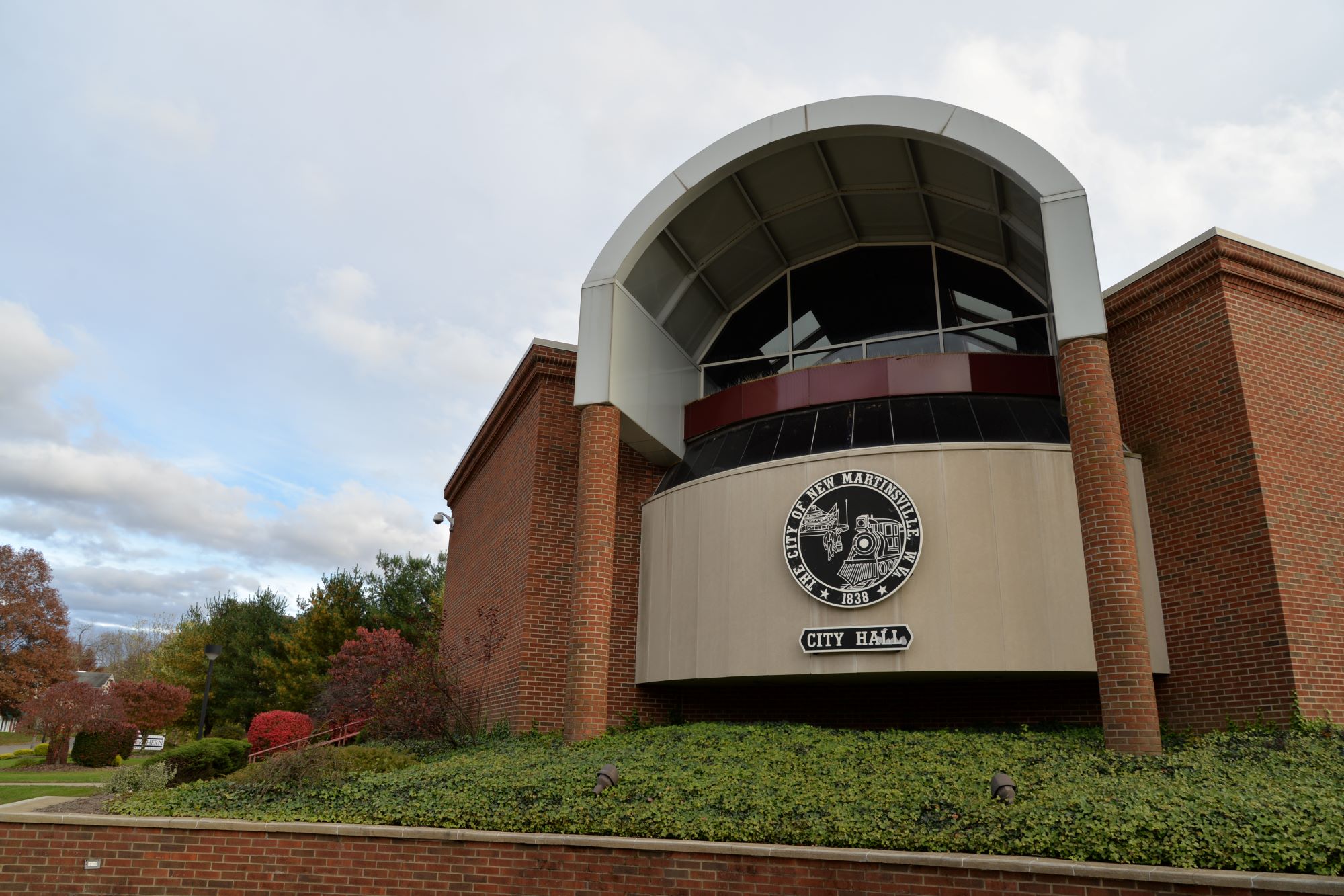 Modern brick city hall building with a prominent arched metal canopy forming a semi-cylindrical cover over the entrance, dark-tinted curved windows beneath it, and a circular city seal centered on a brown facade above landscaped greenery.