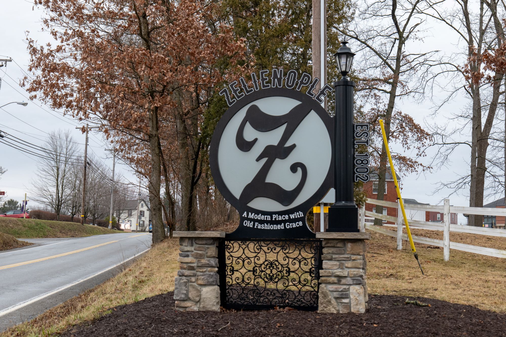 Decorative roadside sign for Zelienople featuring a large stylized “Z,” stone pillars and wrought-iron details, set beside a two-lane road with leafless trees and buildings in the background.