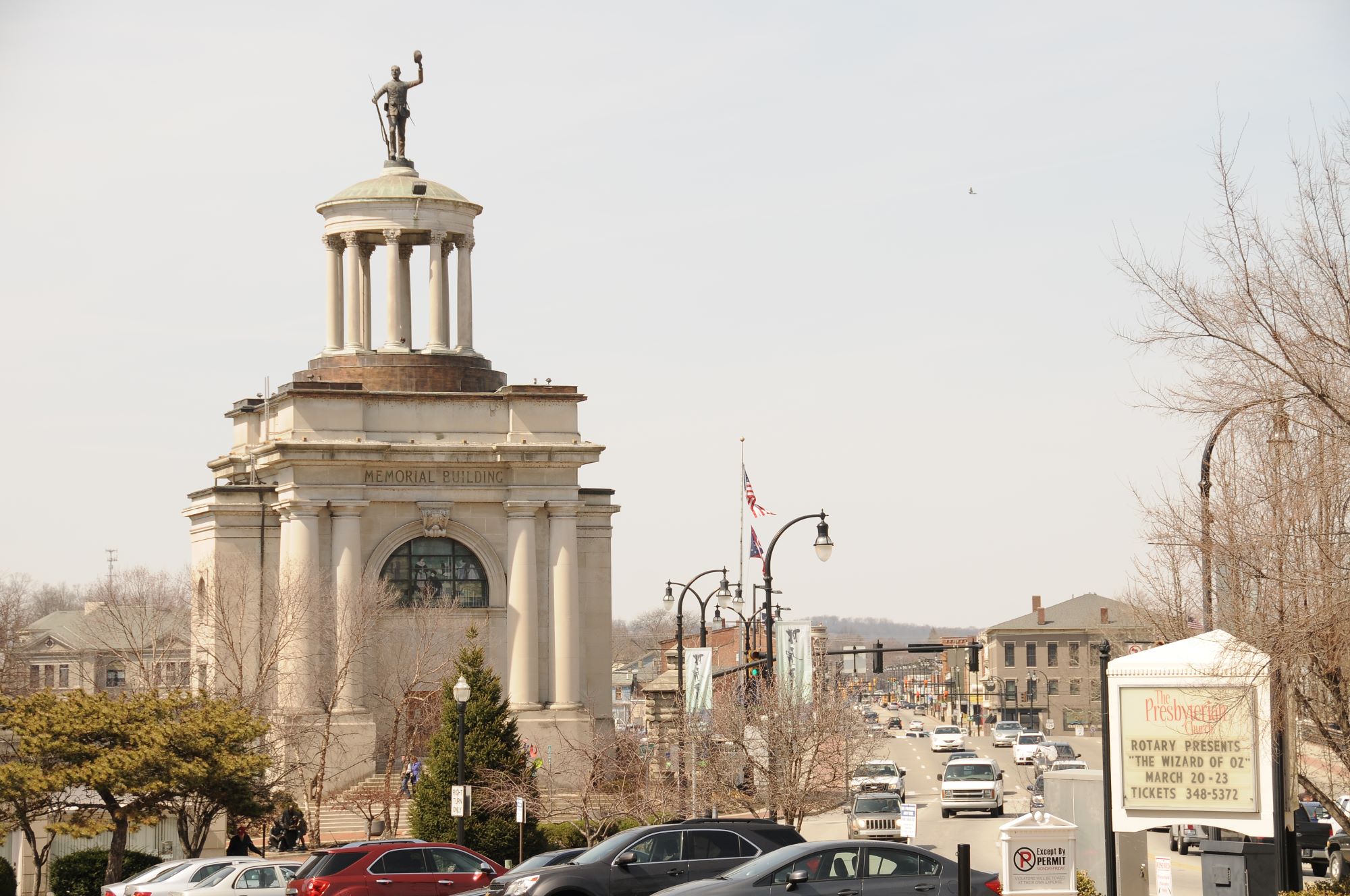 Historic memorial building with a domed rotunda and statue on top overlooks a busy street with cars, storefronts and leafless trees on a clear day.