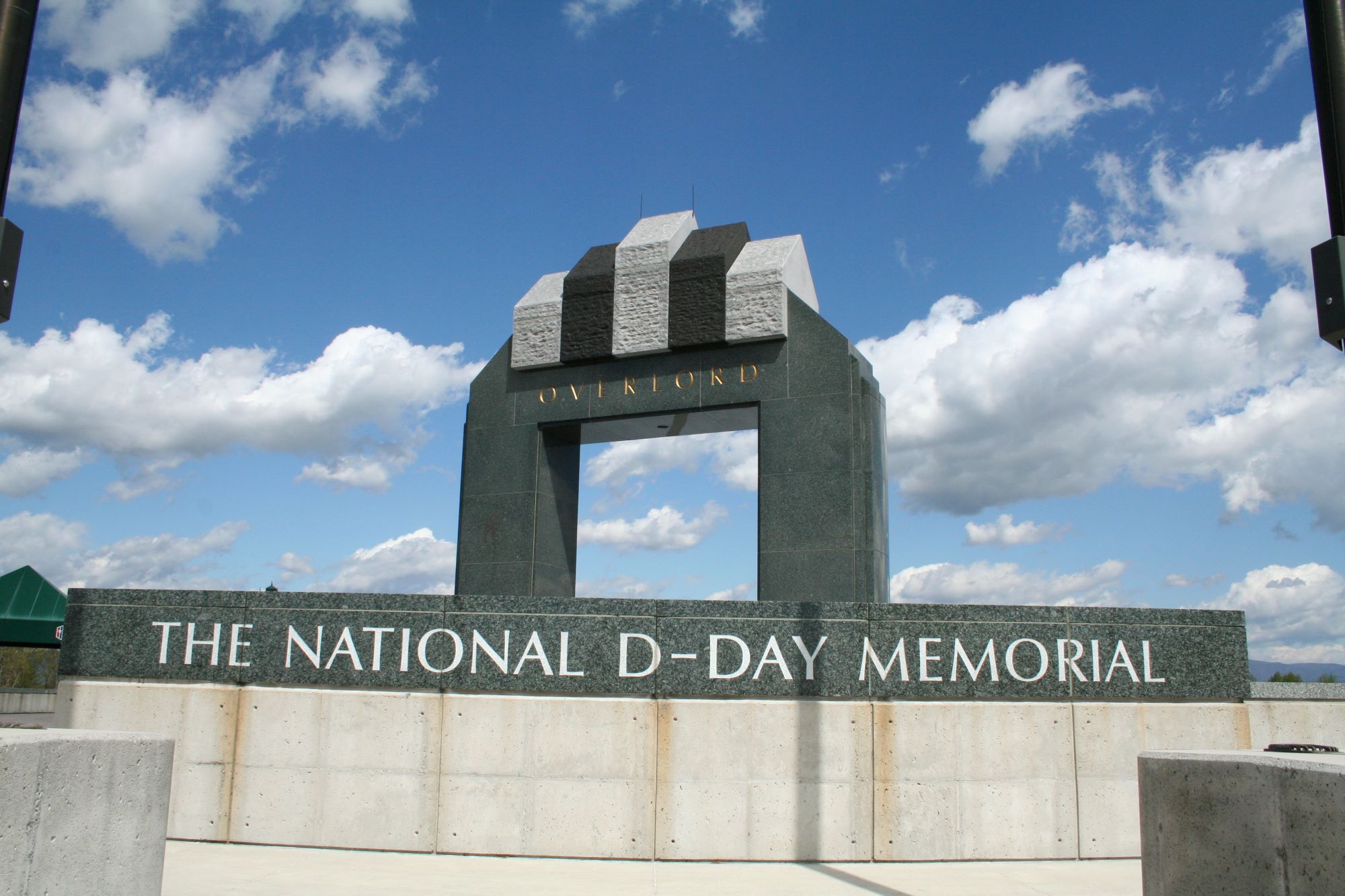 Stone monument at the National D-Day Memorial featuring a large arched structure with sculpted elements above it and engraved lettering on a broad stone wall, set against a blue sky with scattered clouds.