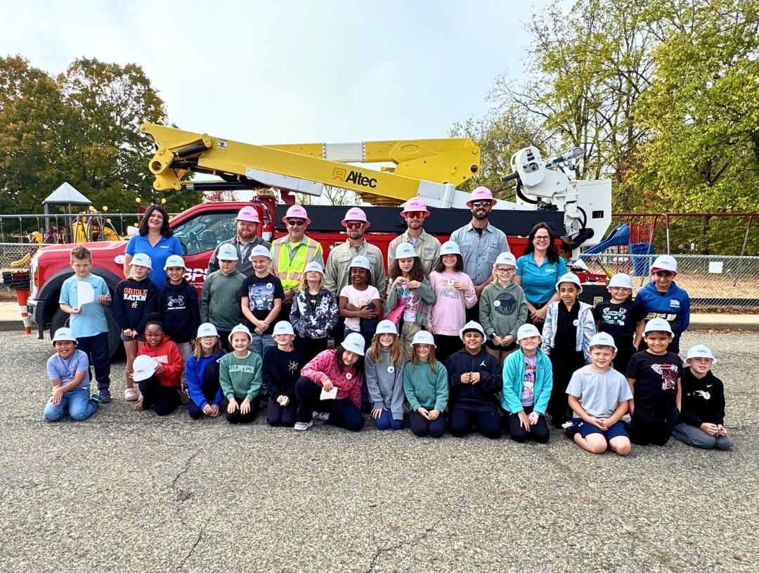 Group of children and adults wearing hard hats pose together outdoors in front of a utility bucket truck and equipment, with a playground and autumn trees in the background.