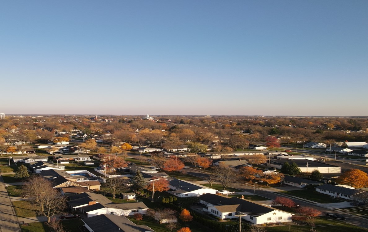 Aerial view of a small town with residential neighborhoods, tree-lined streets and scattered commercial buildings, showing autumn foliage under a clear blue sky.
