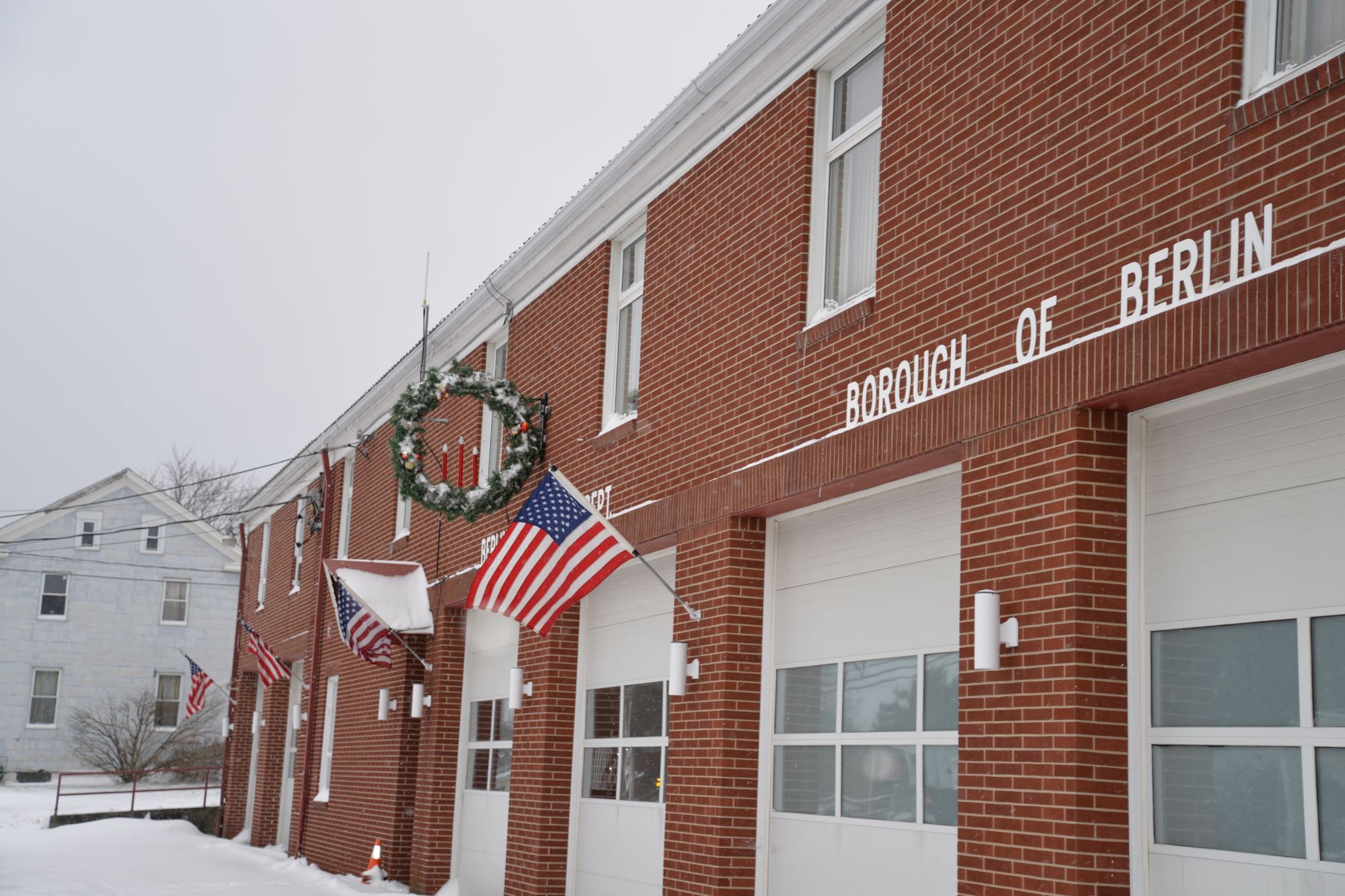 Red brick Borough of Berlin municipal building with garage doors, American flags, and a holiday wreath, set along a snow-covered sidewalk on an overcast winter day.