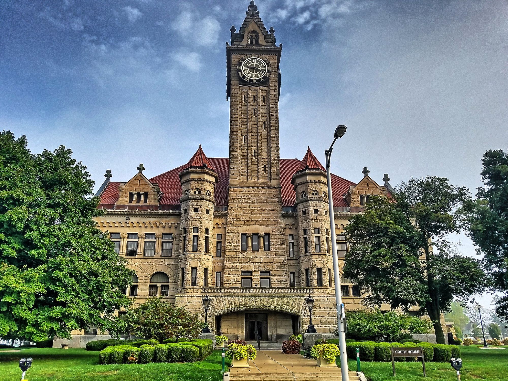 Stone courthouse with a tall central clock tower, red-tiled roof and arched entrance, surrounded by manicured lawns and trees under a partly cloudy sky.