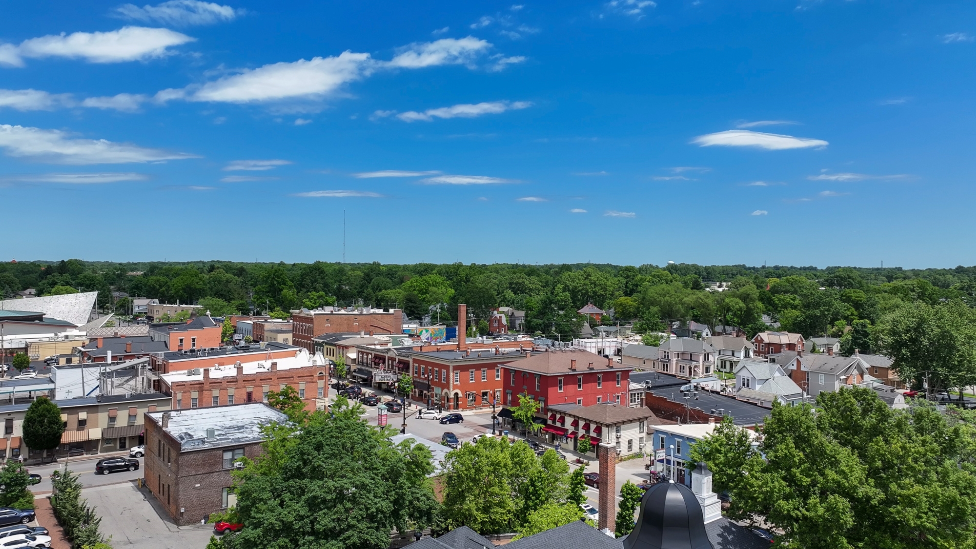 Aerial view of a vibrant downtown district with brick buildings, shops and tree-lined streets, surrounded by dense greenery and residential neighborhoods under a bright blue sky with scattered clouds.