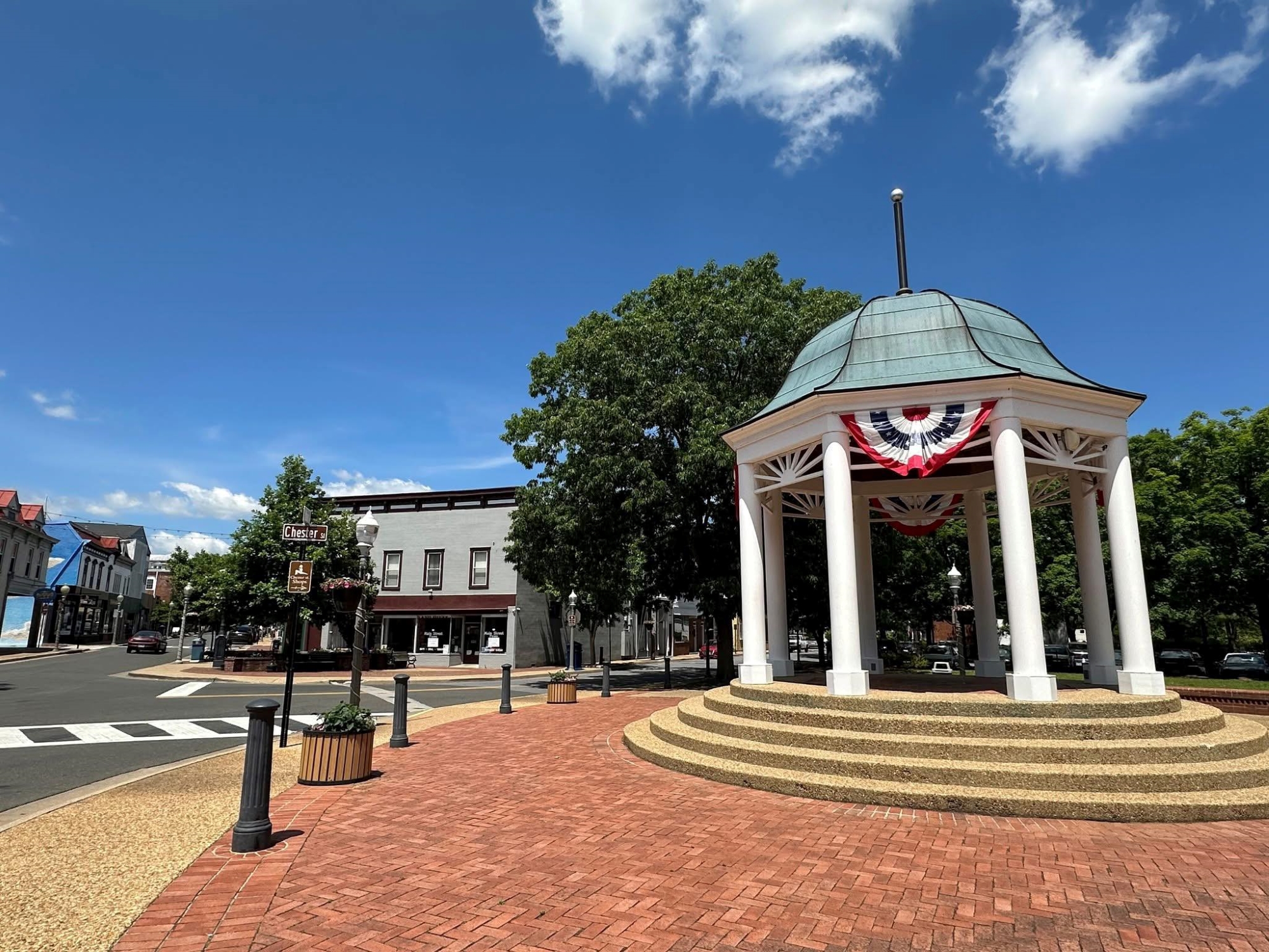A white gazebo decorated with red, white, and blue bunting stands in a small-town square on a sunny day. It sits on a raised circular platform with brick paving around it. Behind the gazebo are trees, a few storefronts, and a street sign reading “Chester St.”
