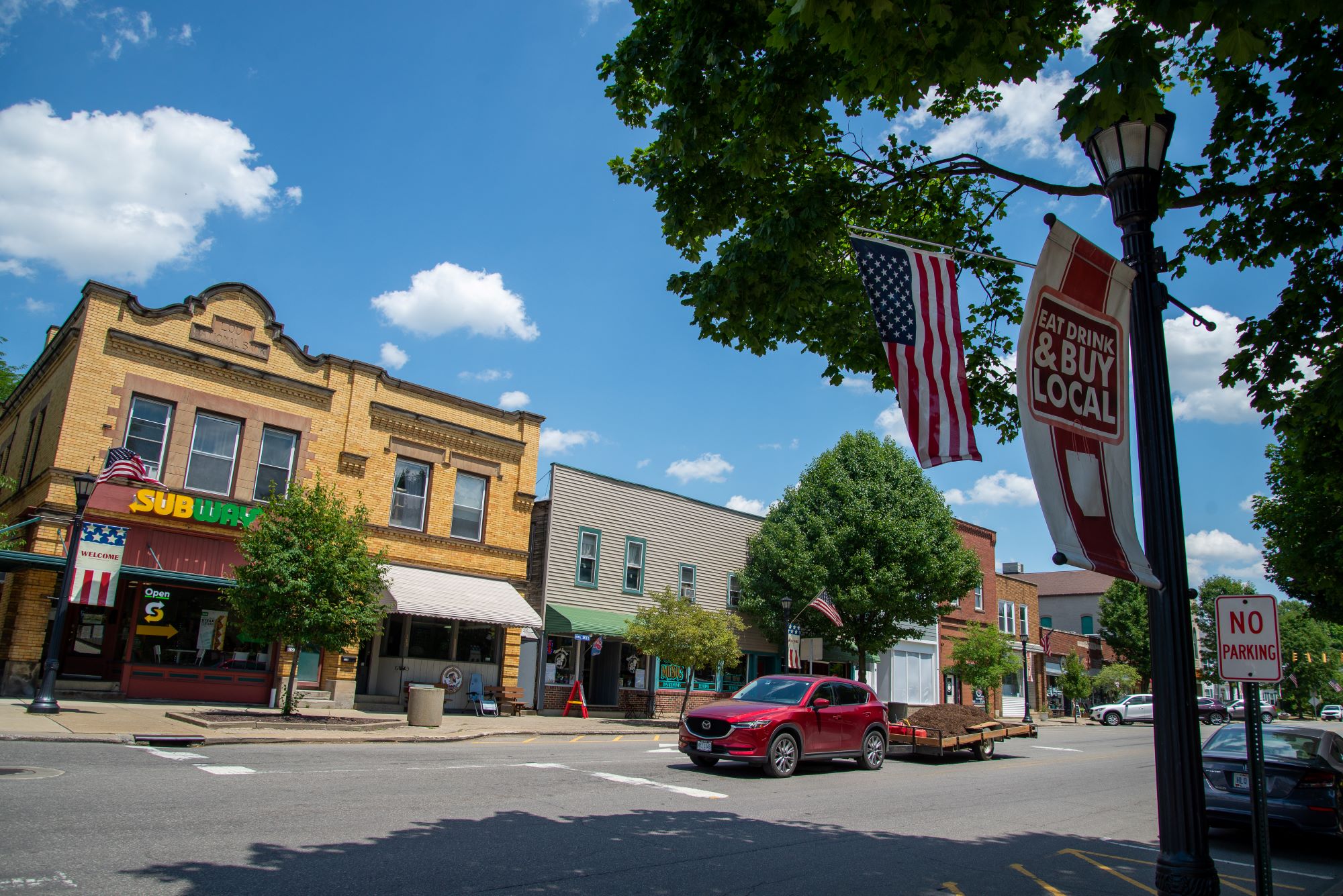 Small-town downtown street with historic brick storefronts, a red SUV driving past, and in the foreground an American flag and a “Eat, Drink & Buy Local” banner hanging from a lamppost beneath leafy trees on a sunny day.