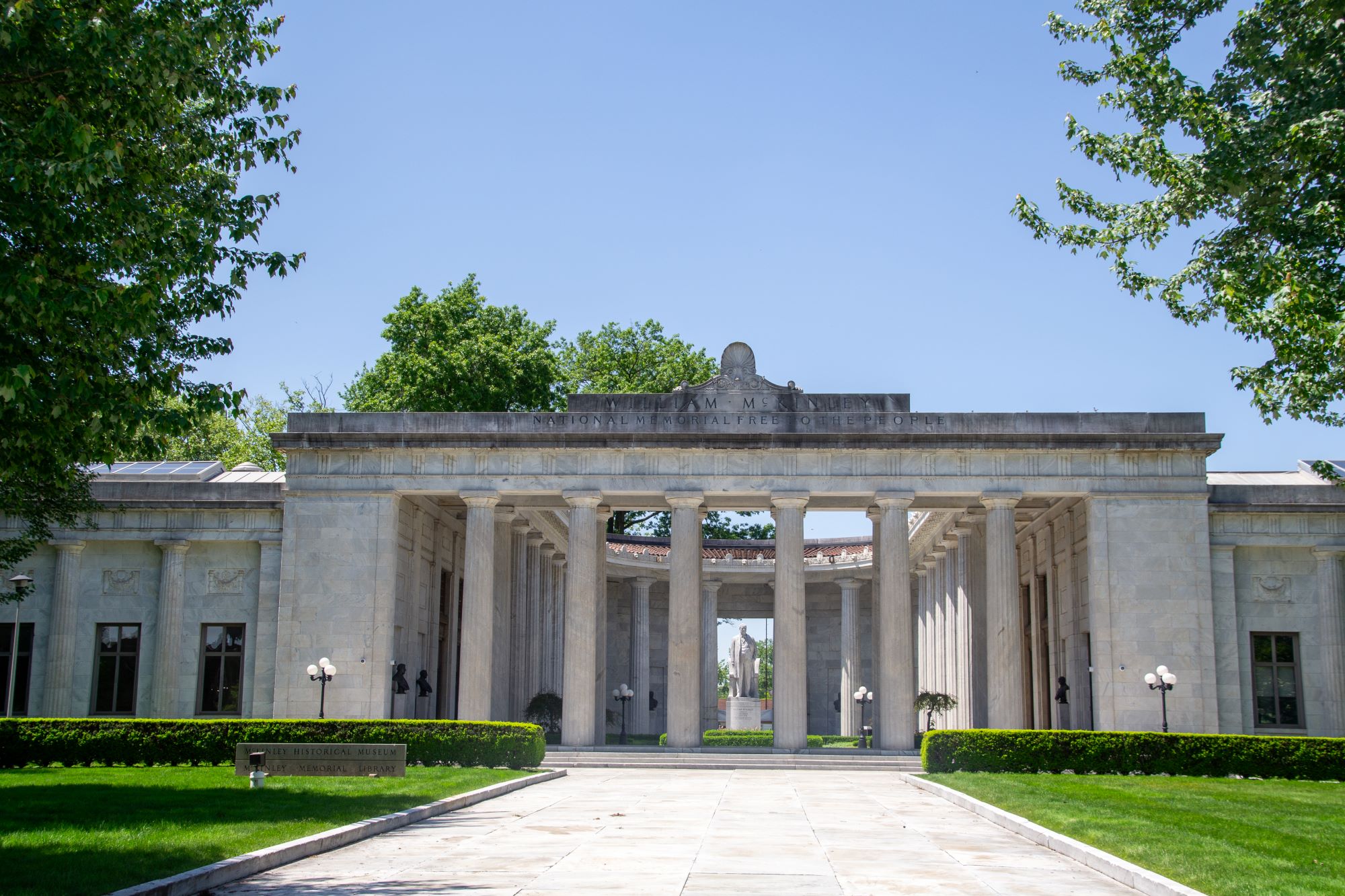Neoclassical memorial with a wide stone colonnade and central open rotunda, flanked by manicured lawns and trees, viewed from a straight walkway under a clear blue sky.