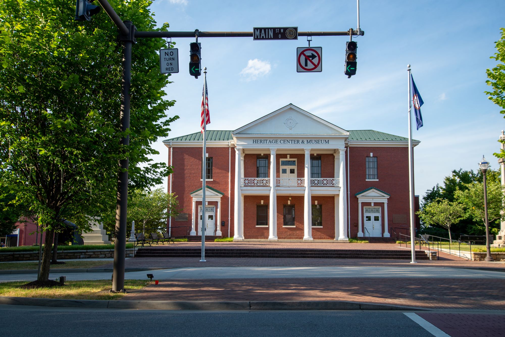 Historic red-brick Heritage Center and Museum with white columns and a green roof, viewed across a street intersection with traffic lights, flags and trees on a sunny day.