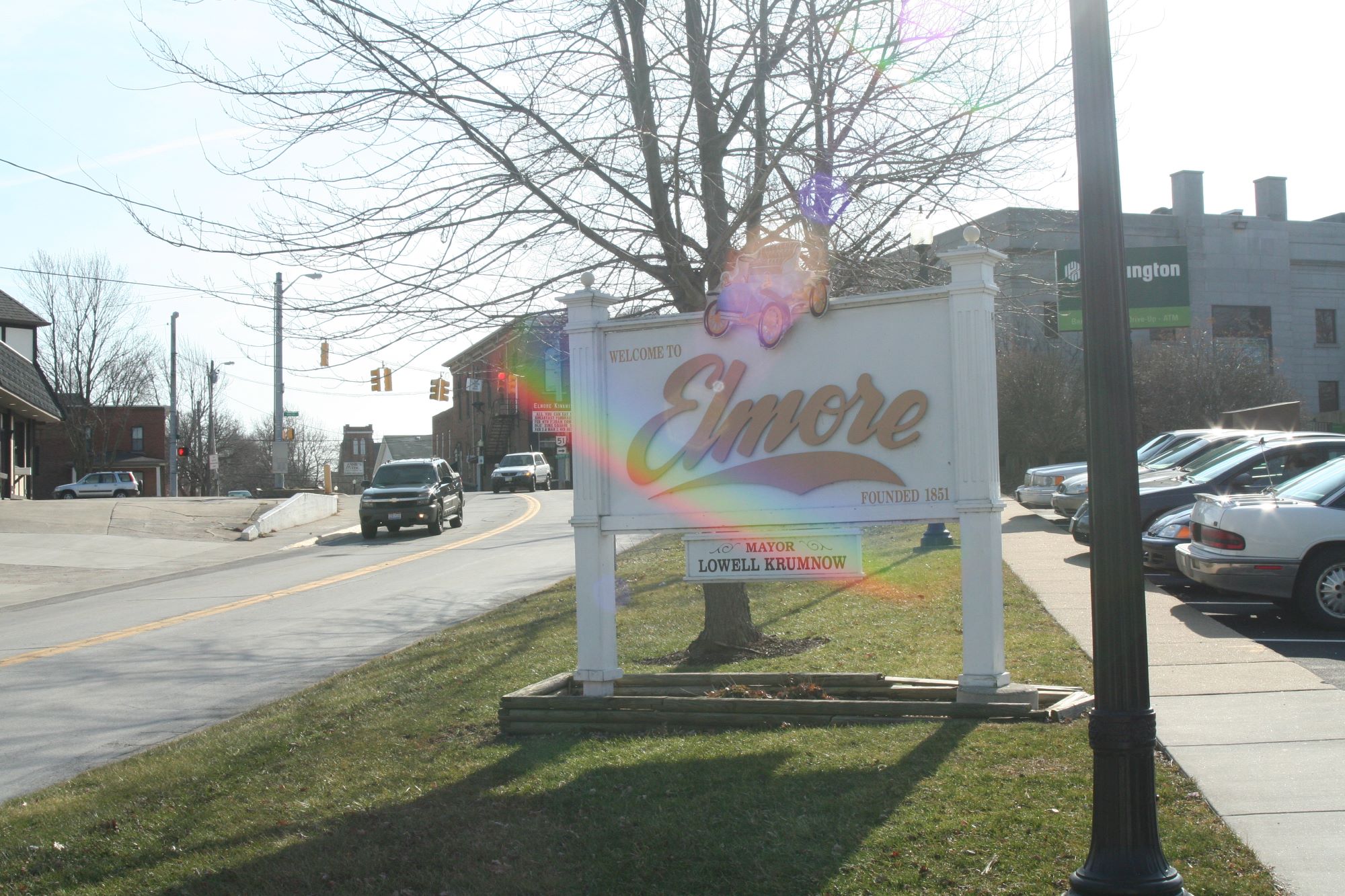 A roadside sign reading “Welcome to Elmore” standing on a grassy area beside a street, with cars, buildings and leafless trees in the background.