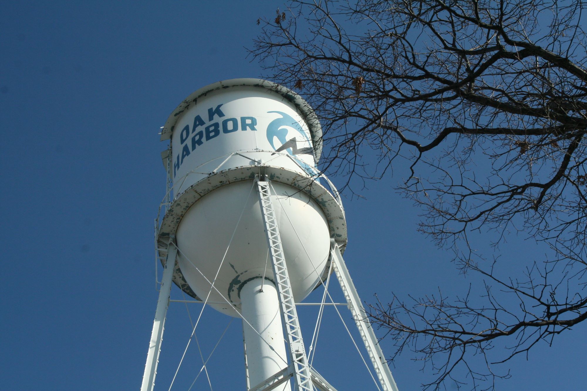 Low-angle view of the Oak Harbor water tower, a white steel structure with blue lettering that says 'Oak Harbor' with a blue power wheel, set against a clear blue sky with bare tree branches in the foreground