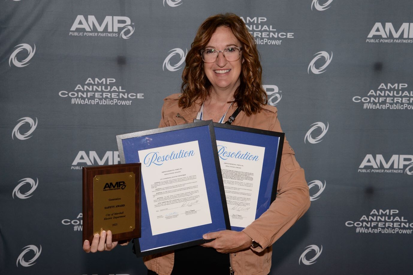Person standing in front of an AMP Annual Conference backdrop holding a wooden award plaque and two framed resolutions.