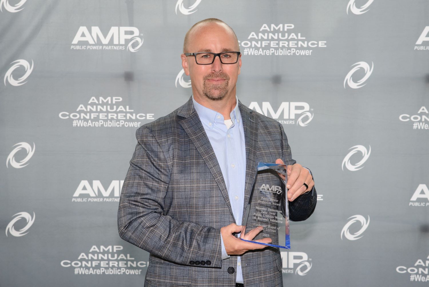 Person standing in front of an AMP Annual Conference backdrop, holding a clear glass AMP award with both hands.