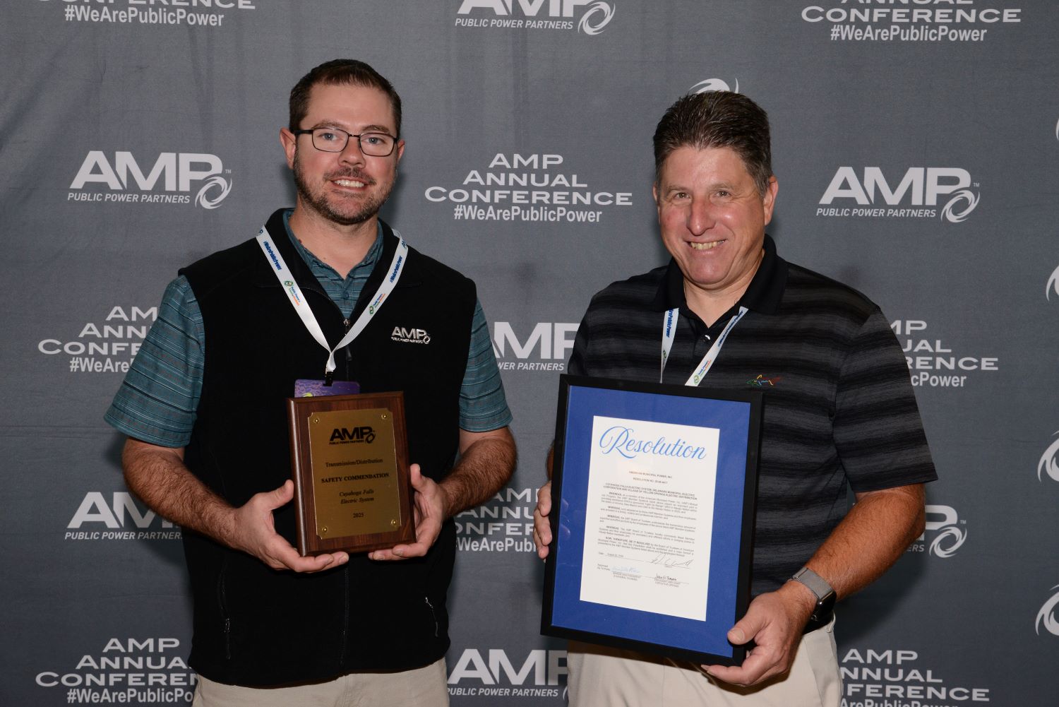 Two people standing side by side in front of an AMP Annual Conference backdrop. The person on the left holds a wooden AMP plaque award, and the person on the right holds a framed resolution document with a blue border. Both are wearing conference lanyards, and the backdrop displays AMP Public Power Partners logos and the text ‘#WeArePublicPower.'