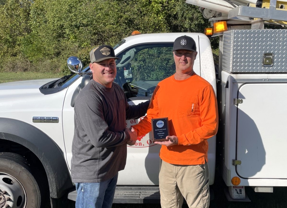 Two utility workers stand beside a white service truck, shaking hands while one holds a small recognition plaque. The truck has equipment compartments and a ladder on top, and trees are visible in the background on a sunny day.