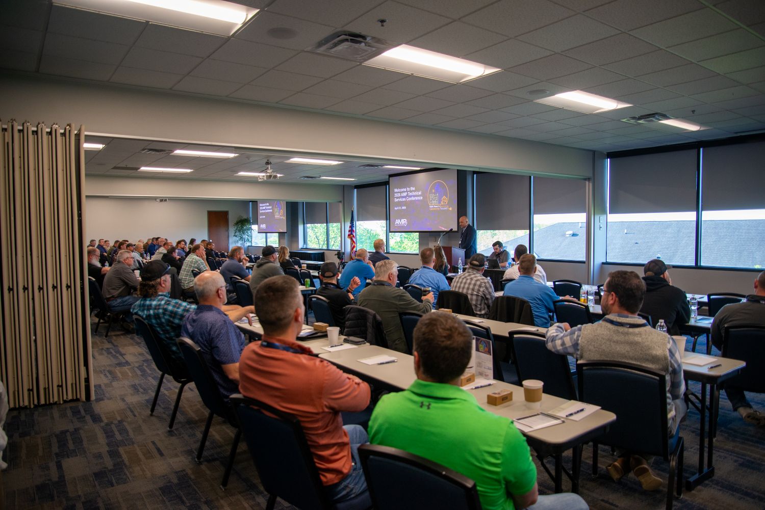 Attendees seated at tables listening to a speaker presenting at the front of a conference room during the AMP Technical Services Conference, with presentation slides displayed on large screens.