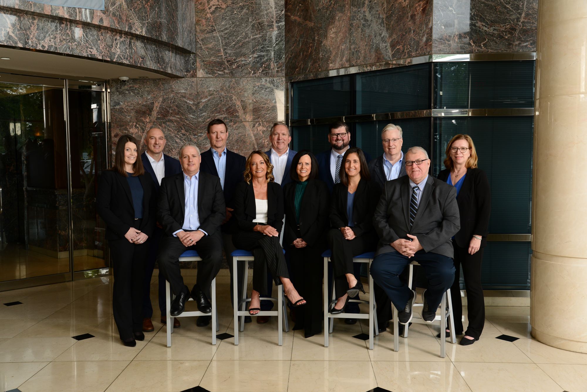 A group of twelve people, dressed in business attire, are posing for a formal photograph in an indoor setting with marble walls and a tiled floor. Seven individuals are standing at the back while five are seated on stools in front.