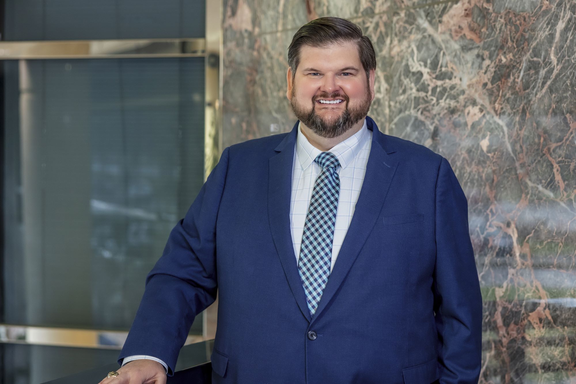 A person wearing a navy-blue suit, a white dress shirt with a subtle grid pattern, and a blue patterned tie stands indoors beside a dark countertop. A polished stone wall with mixed gray and pink tones is in the background.