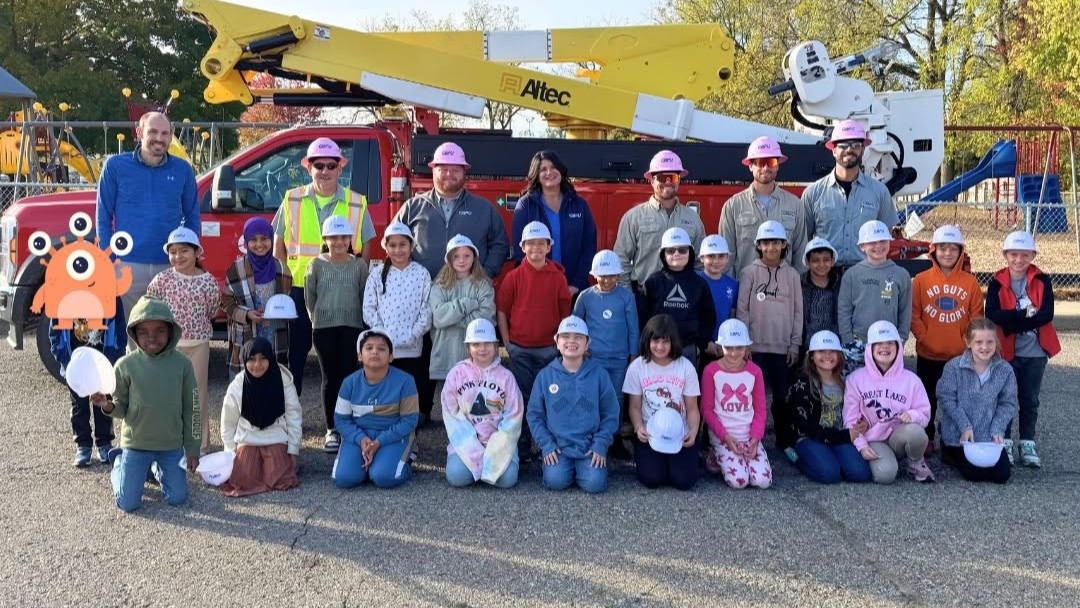 A group of elementary school students and Coldwater Board of Public Utilities employees pose in front of a utility bucket truck during an educational community outreach event. The students are wearing CBPU-branded hard hats, highlighting the utility's efforts to engage local youth and connect the community with public power services.