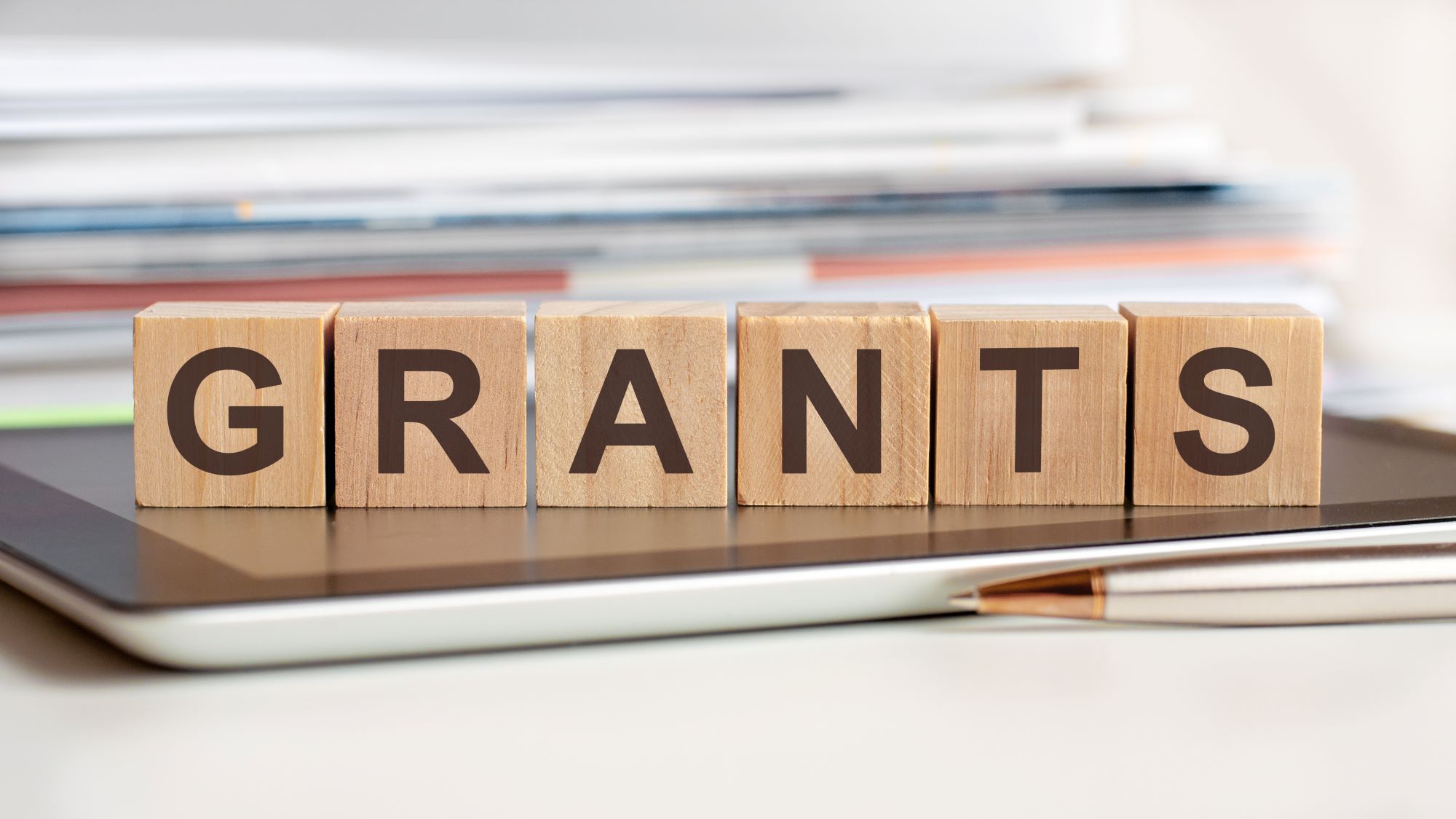 Wooden letter blocks spelling “GRANTS” sit on a tablet, with a pen in the foreground and stacked documents in the background