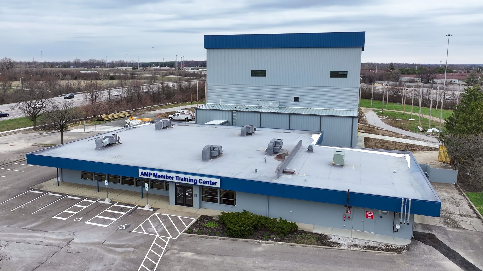 Aerial view of the AMP Member Training Center building with a flat roof, rooftop HVAC units, blue trim, and a marked parking lot in front and roadway in the background.