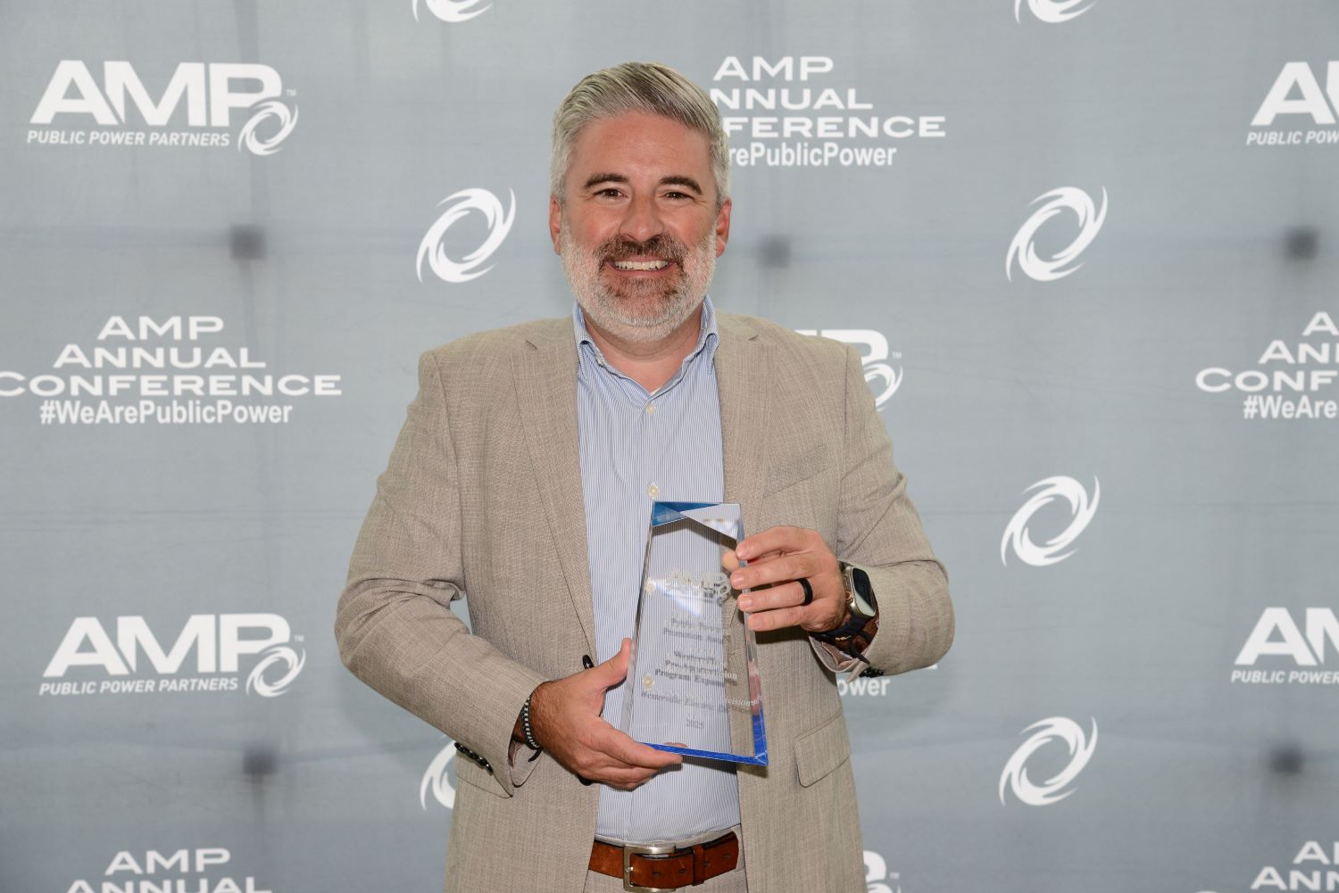 Person standing in front of an AMP Annual Conference backdrop, holding a clear glass AMP award with both hands.