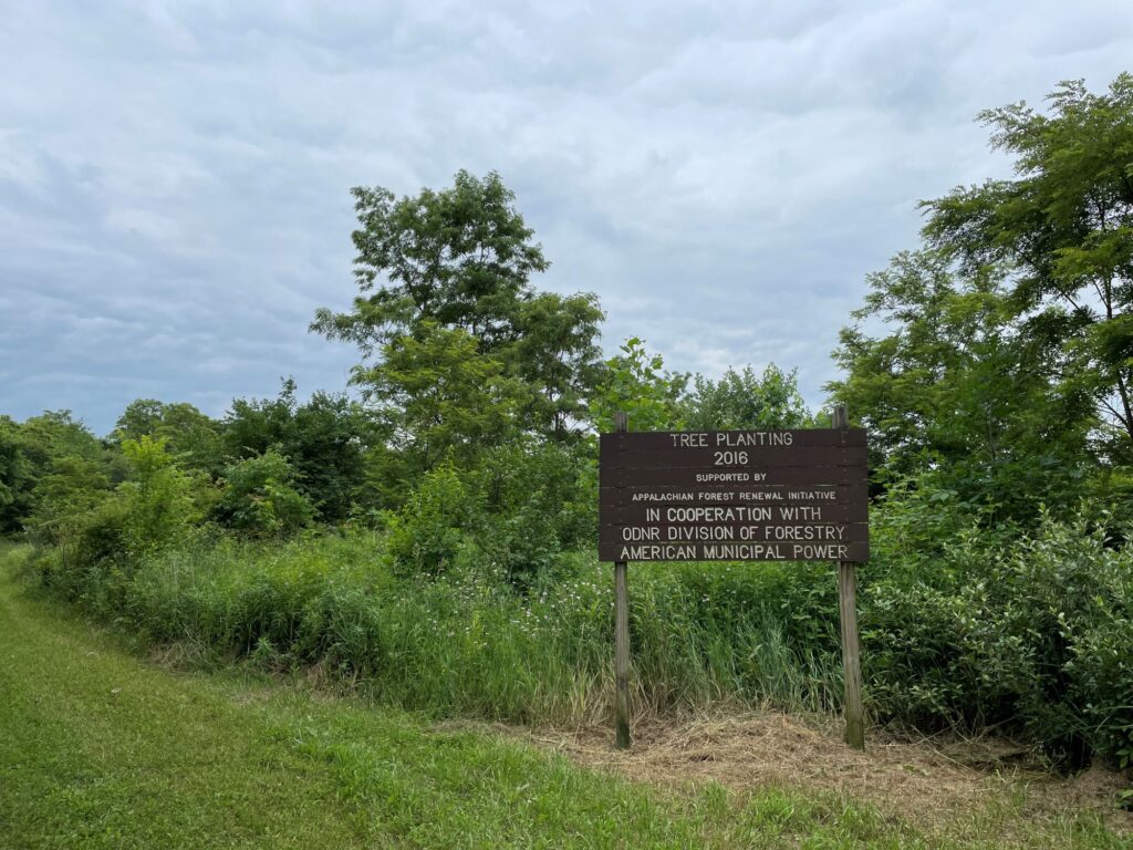 Wooden sign reading “Tree Planting 2016,” supported by Appalachian Forest Renewal Initiative in cooperation with ODNR Division of Forestry and American Municipal Power, stands in a grassy clearing surrounded by dense green trees under an overcast sky.