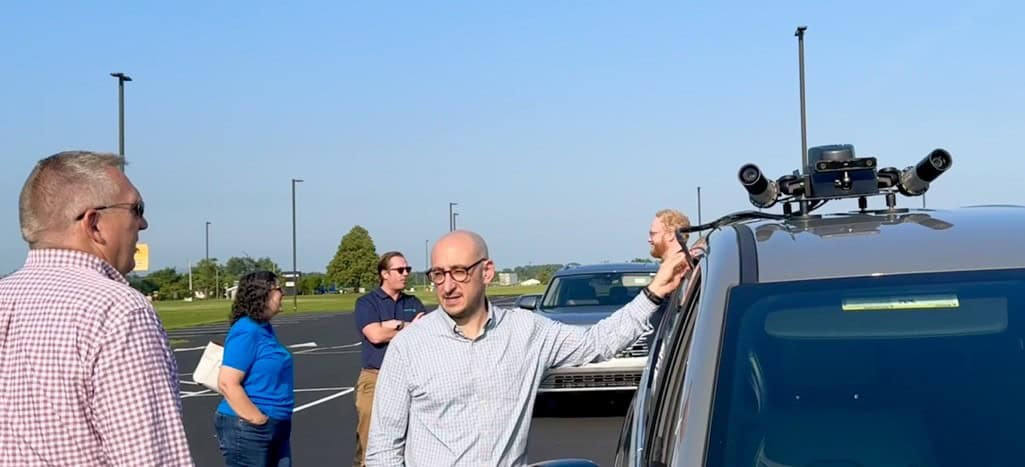 Several people stand in a parking lot beside a vehicle equipped with a roof-mounted, Noteworthy AI camera