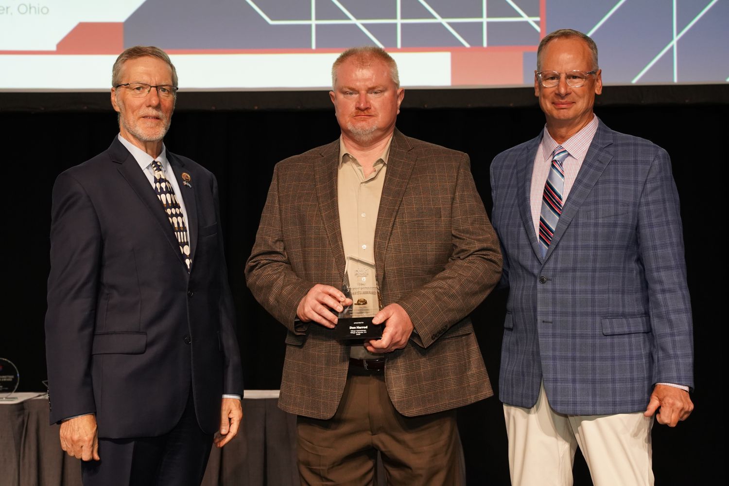 Three individuals stand on a stage during an awards ceremony, with the center individual holding a glass trophy. The award recognizes Don Harrod with the APPA Seven Hats Award. The two individuals on either side wear business suits, and a large presentation screen with graphic elements and partial text is visible in the background.