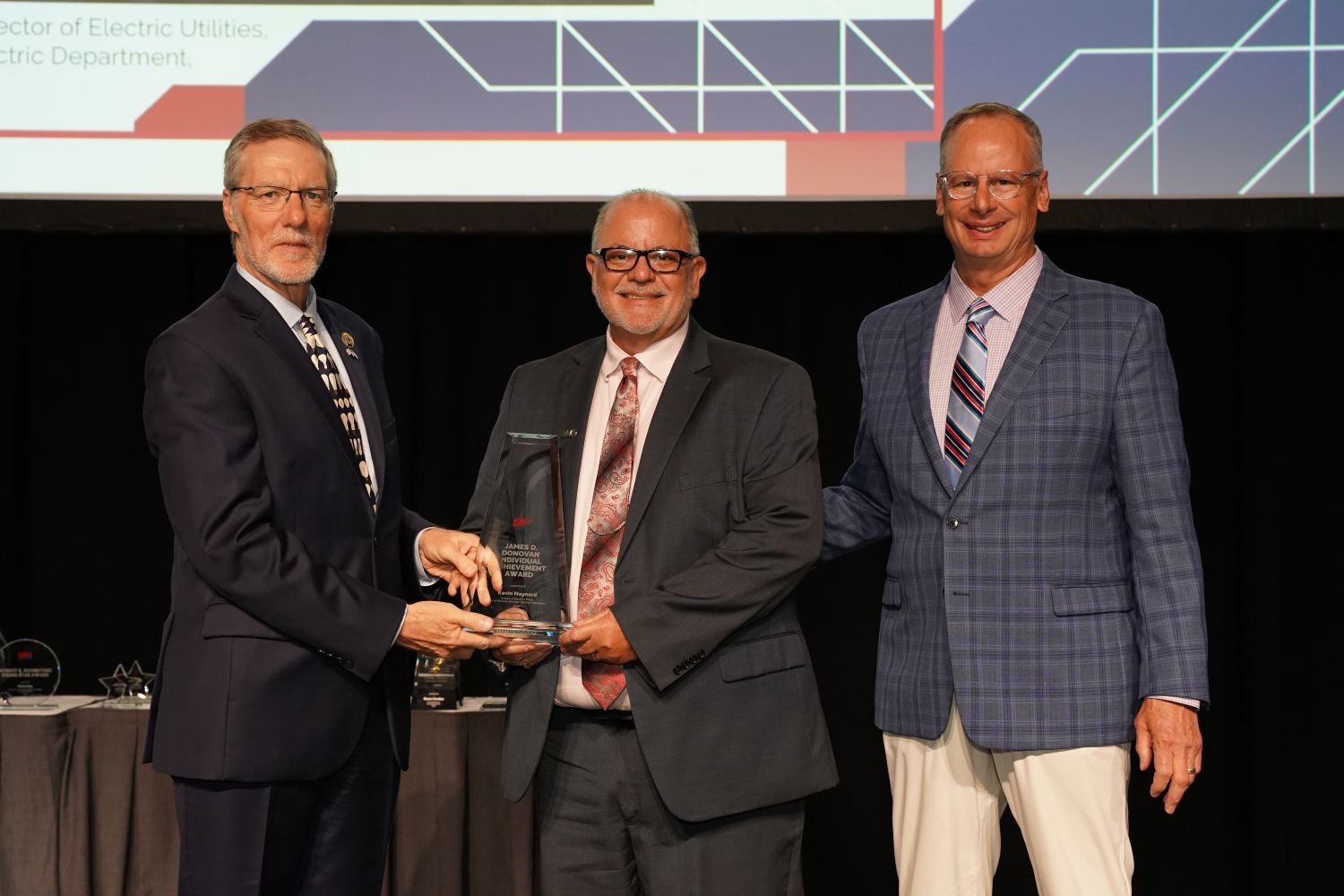 Three individuals stand on a stage during an awards ceremony while a glass award is presented to Kevin Maynard for the James D. Donovan Individual Achievement Award. The recipient holds the award as another individual presents it and a third stands beside them. All are dressed in business attire, with a large presentation screen displaying graphic elements in the background.