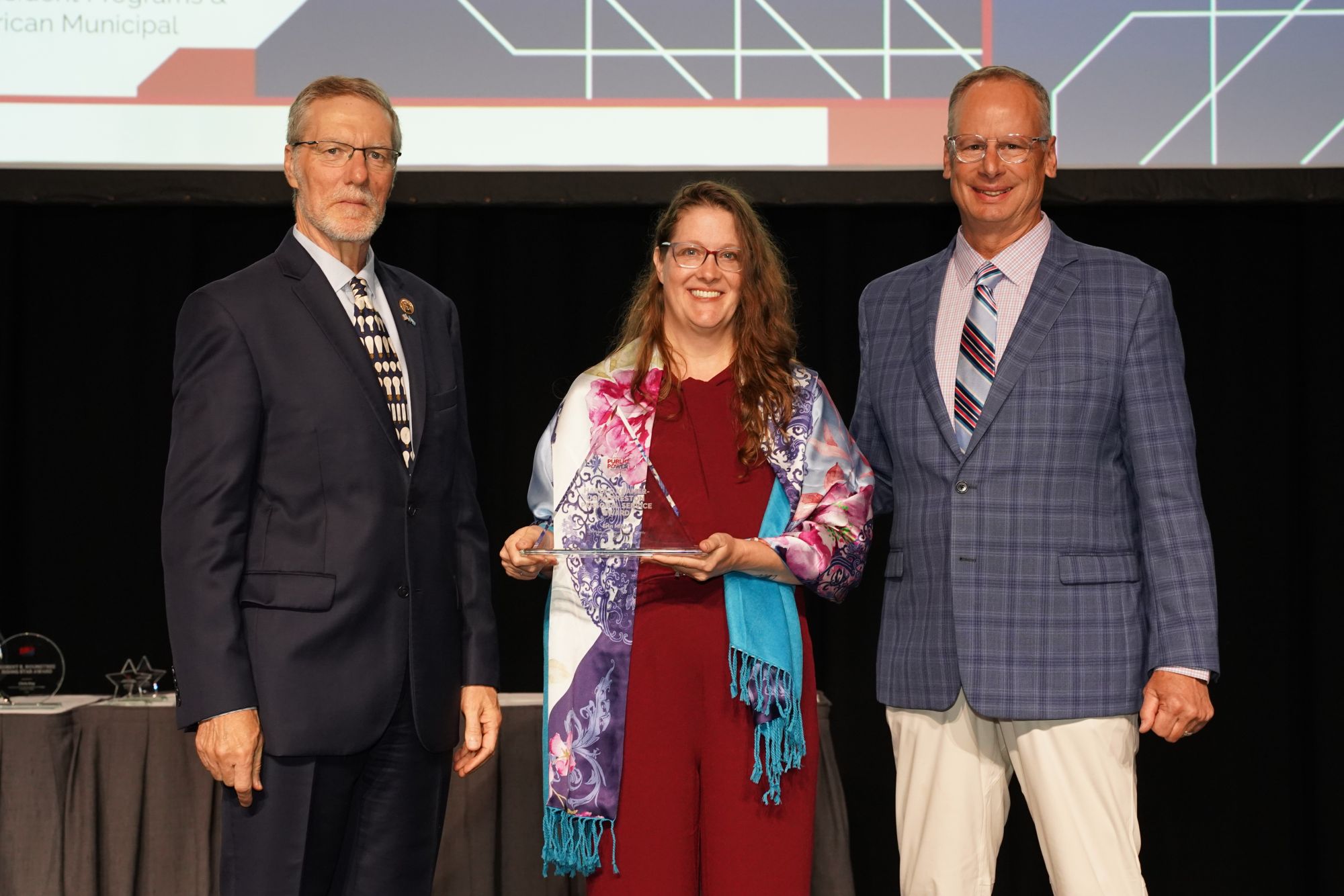 Three individuals stand on a stage at an awards ceremony as the center individual receives a glass award. The award recognizes Erin Miller with a Public Power Service Award. The presenter on one side hands over the award while another individual stands alongside. All are dressed in business attire, with a large presentation screen displaying graphic elements in the background.