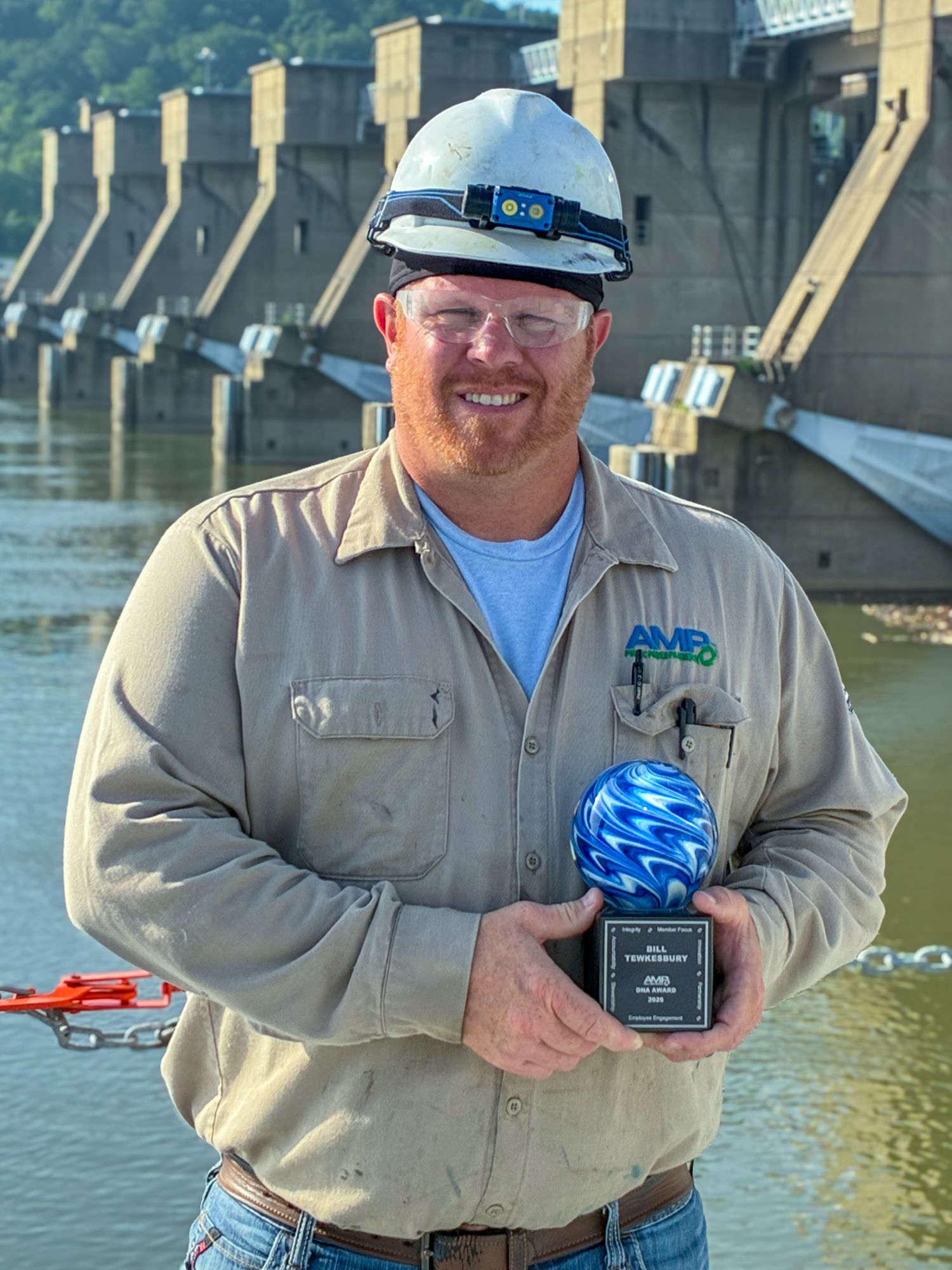 Bill Tewkesbury, Plant Operator at AMP’s Willow Island Hydroelectric Plant and recipient of the 2026 AMP DNA Award, stands in front of the hydroelectric facility holding his award. He is wearing a hard hat, safety glasses and an AMP work shirt, with the dam and water visible in the background.